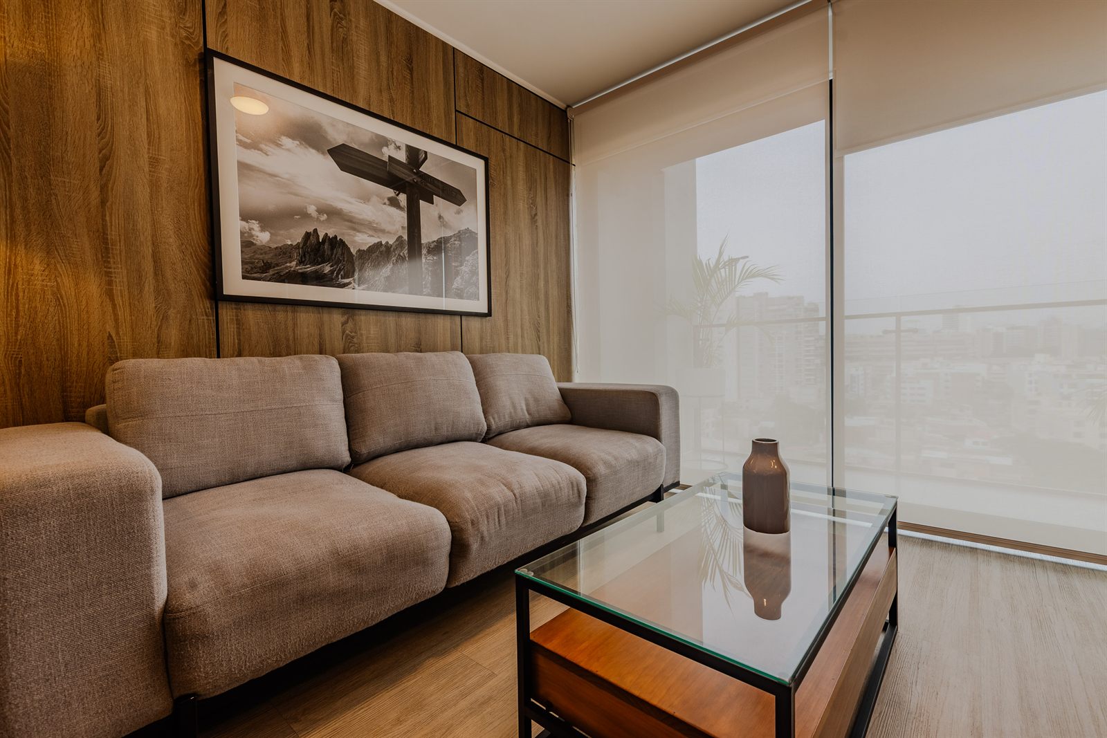 Sala de un departamento en Barranco con pared de madera, sofá de tela beige y ventanal con vista a la ciudad