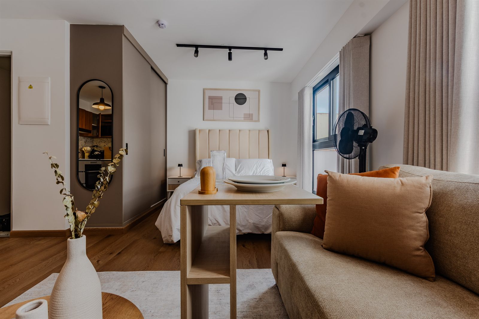 Estudio en Miraflores con cama de cabecera beige, sofá con cojines naranjas y mesa de comedor de madera clara
